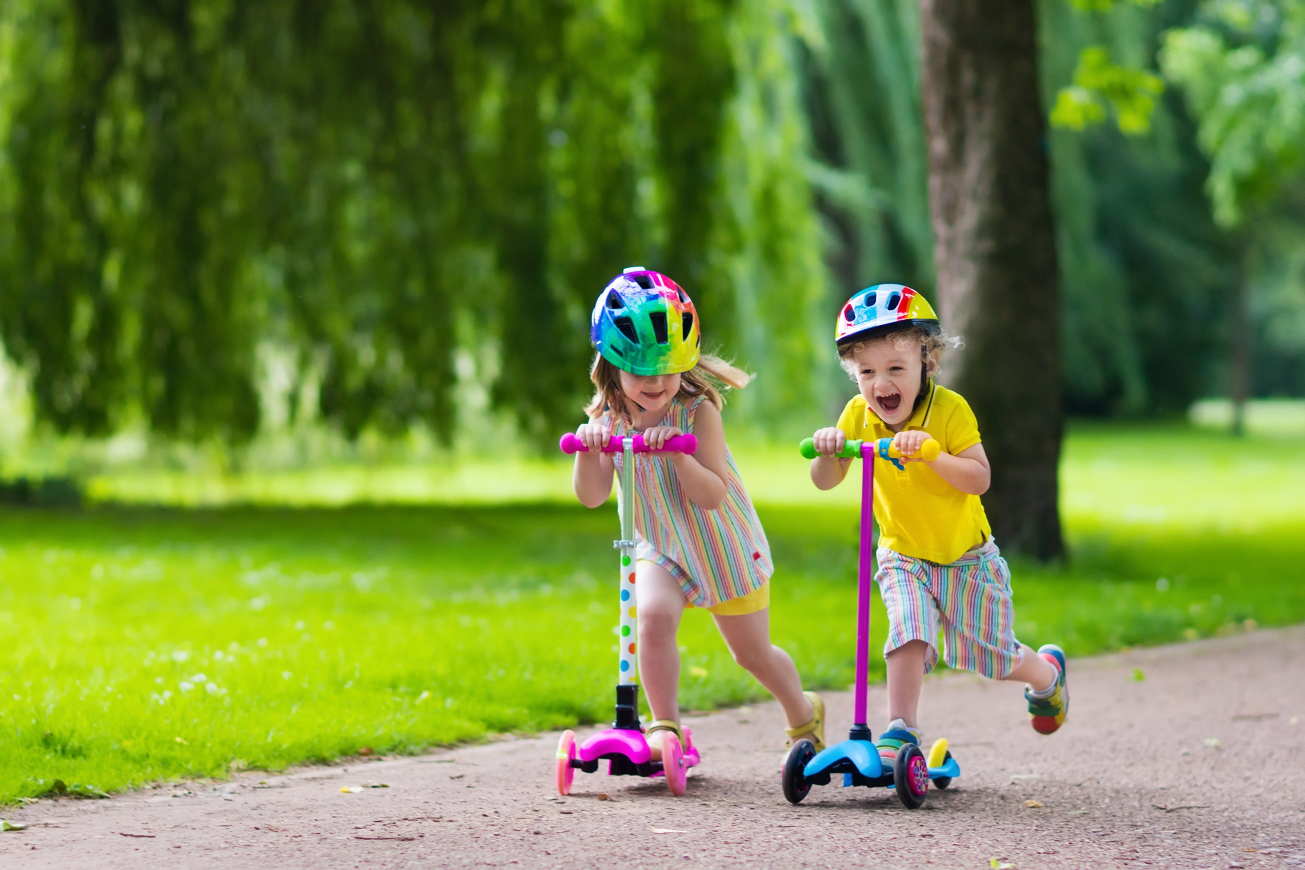 Children wearing helmets ride scooters through a park